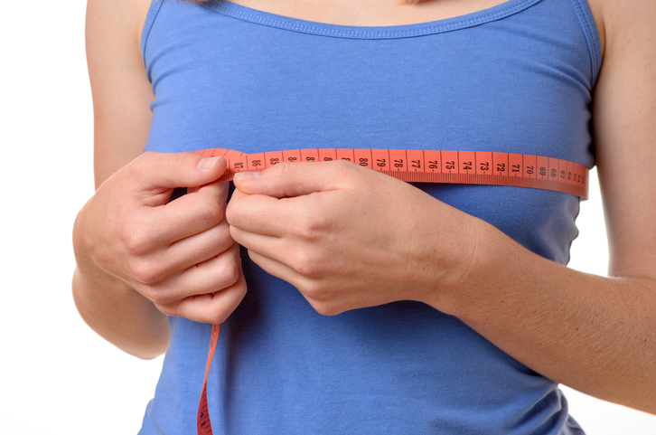 Young woman wearing a blue summer t-shirt measuring her breasts with a tape measure, close up torso view on white