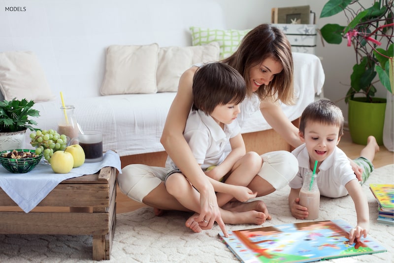 Woman sitting on the floor with her two young sons, looking at an illustrated book cover.