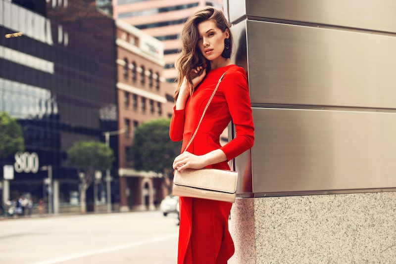 Thin, beautiful woman wearing a red dress and leaning against a building.